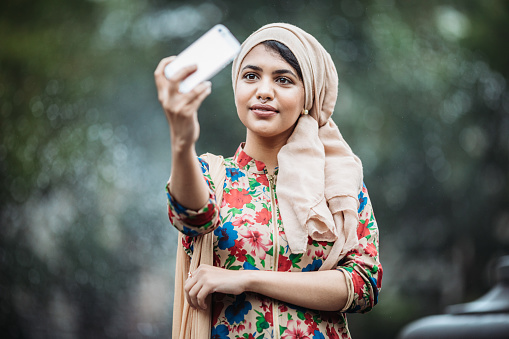 A beautiful young woman from Bangladesh spends some leisure time exploring New York city.  She takes a selfie with her smartphone.  She wears a hijab and traditional clothing.