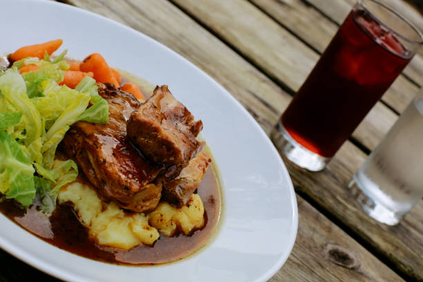 Lamb shank with vegetables on an pub outdoor wooden table, glass of water and juice next to it
