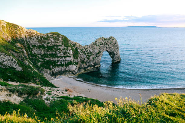 View from the hill over people walking on a beach by the sea and Durdle Door, UK View from the hill over people walking on a beach by the sea and Durdle Door, a natural limestone arch on Dorset's Jurassic Coastline, UK durdle door stock pictures, royalty-free photos & images