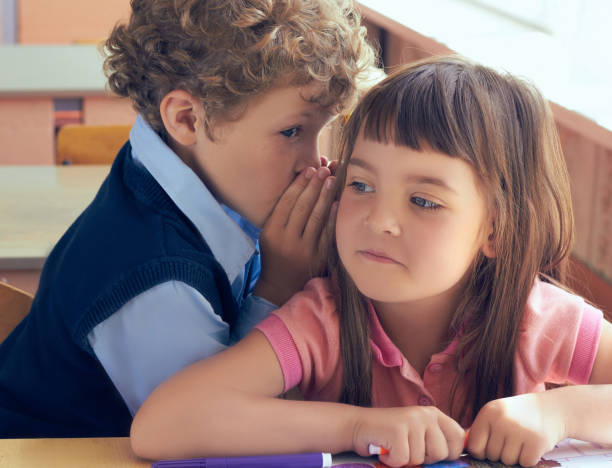 istock education, elementary school, learning and people concept - smiling schoolboy whispering secret to classmate ear in classroom 865967396