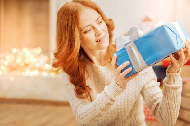 Surprised mature lady looking at christmas present Have no idea what is inside. Amazed redhead woman wearing a warm knitted sweater sitting on the floor and looking at a beautifully wrapped gift with excitement. hair covering eyes stock pictures, royalty-free photos & images