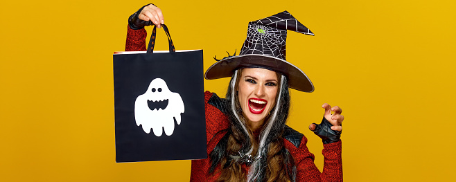 Colorful halloween. smiling modern woman in halloween witch costume isolated on yellow background showing shopping bag and frightening
