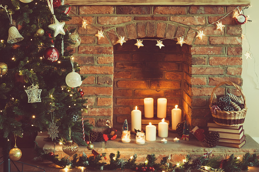 Decorated Christmas tree in front of the fireplace with various candles, pine cones and baubles, selective focus; dark vintage style toned photo