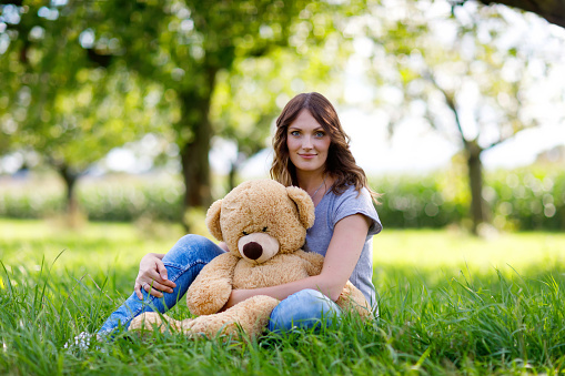 Beautiful young woman sitting on green grass in summer. Lady with long hairs holding huge teddy bear.