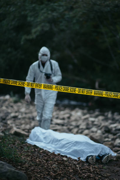 Forensic scientist holding a camera and going towards the dead body A forensic scientist is seen holding a camera and going towards the dead body that is lying on the ground near rocks. The cordon tape is in the middle of a picture. people covered in mud stock pictures, royalty-free photos & images