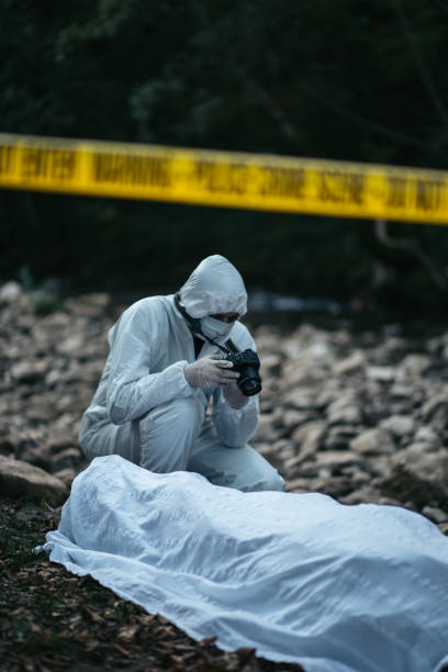 Forensic scientist crouching near the dead body and photographing it A forensic scientist is seen crouching near the dead body while photographing it in the woods by the rocks. The cordon tape is in the middle of the picture. people covered in mud stock pictures, royalty-free photos & images
