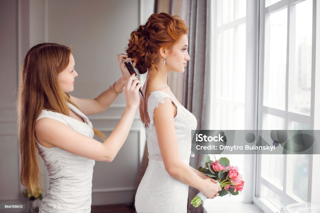 Bride's hairstyle preparation Master stylist makes the bride wedding hairstyle using hairbrush indoors at home near window. Beautiful bride perfect style. Wedding hairstyle make-up luxury wedding dress. Bride Stock Photo