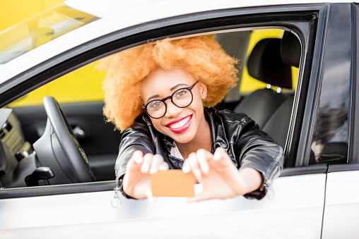 Portrait of a beautiful smiling african woman in leather jacket showing a card with blank space sitting in the car on the yellow background