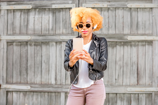 Lifestyle portrait of an african woman in leather jacket making selfie photo with phone standing on the wooden wall background