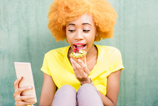 Portrait of a pretty african woman in yellow t-shirt making selfie photo with sweet dessert on the green background