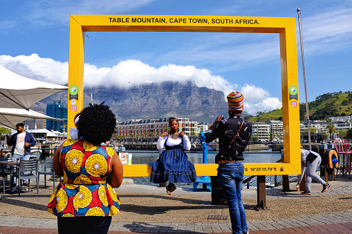 Cape Town,South Africa - September 14, 2017: A young African woman is sitting on the yellow Nature's New 7 Wonders picture frame with the famous Table Mountain in the background. A man is taking  pictures . 