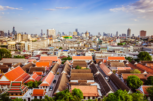 Spectacular view on the capital city of Thailand, under clear blue sky and small white clouds, private houses and buddha temples with red roofs, green trees and tall skyscrapers