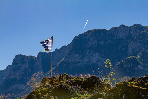 Greek flag and mountains above the Vikos gorge in Epirus, Greece with aircraft contrails in the distance