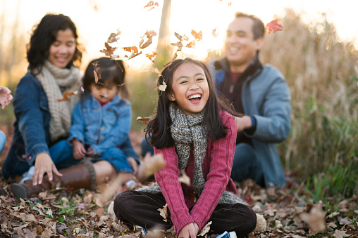 An Ethnic family is outdoors in a field on an autumn day. They are wearing warm clothes. They are sitting in leaves and the daughter has thrown them in the air.