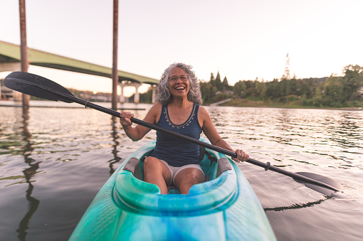 Ethnic female senior citizen kayaks in river on a summer evening. She has a huge smile. There is a bridge behind her.