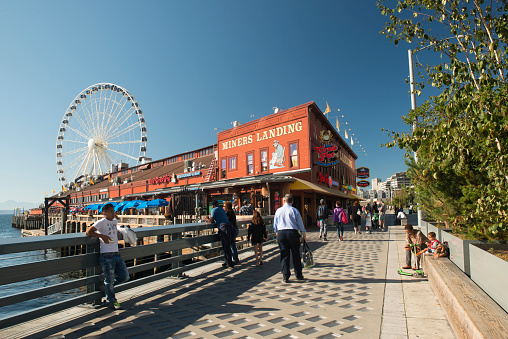 Seattle, USA - September 27, 2017. Tourists on waterfront walk at Pier 57 on Alaskan Way in downtown Seattle with view of Miners Landing food court building and Ferris Wheel (Seattle Great Wheel), Washington State.