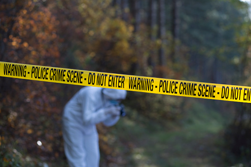 Crime scene in the woods with the focus on the yellow cordon tape and forensic taking the picture