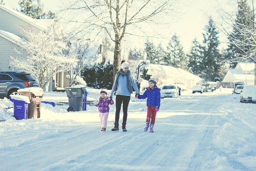 Mom walks with her two young daughters down the snowy neighborhood street. The streets, cars, and sidewalks are covered with snow. They are holding and walking in the middle of the street with big smiles on their faces.
