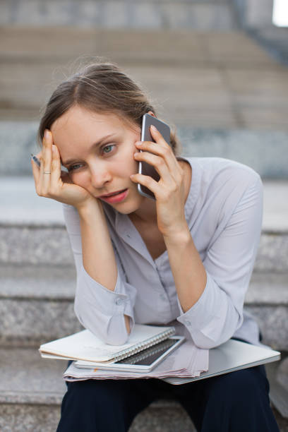 Portrait of tired young Caucasian businesswoman sitting on stairs with notepads and calling up client. Overload concept