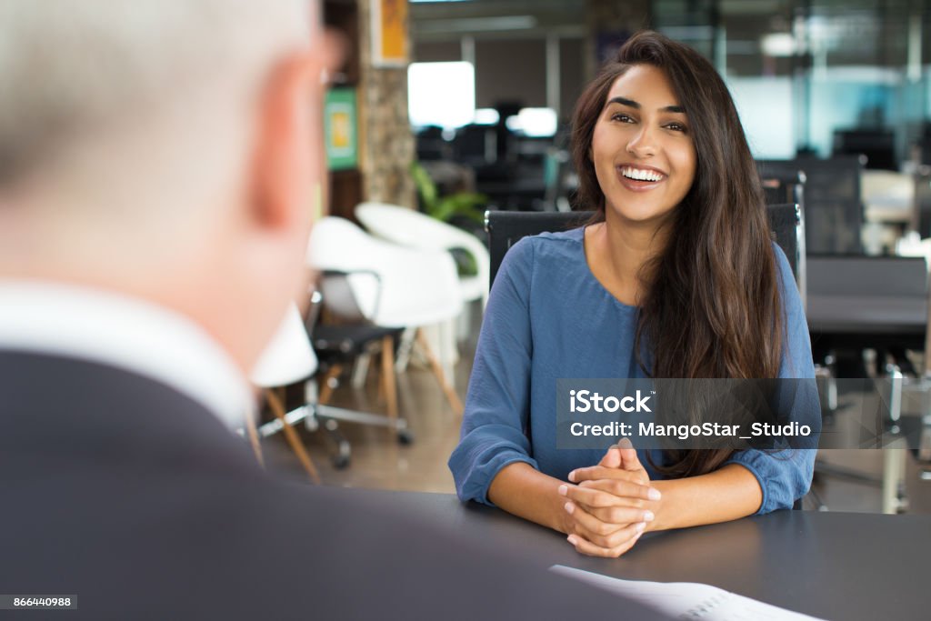 Young female candidate laughing at job interview Portrait of young Indian female candidate sitting at table opposite senior executive and laughing at job interview. Recruitment concept Job Interview Stock Photo