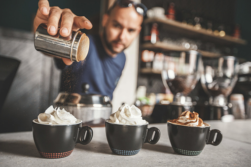 Barista is spreading cinnamon on a coffee with whipped cream. Except the simple coffee with whipped cream, he is making coffee with extra cinnamon and cocoa powder.
