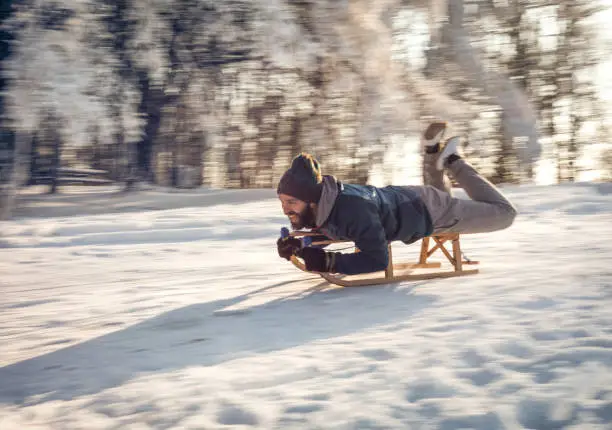 Playful man having fun while moving down with a sledge in winter park. Blurred motion.