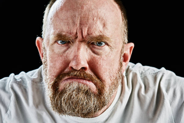 A mature bearded man looks at camera, grimacing in disapproval, looking threatening and aggressive.
