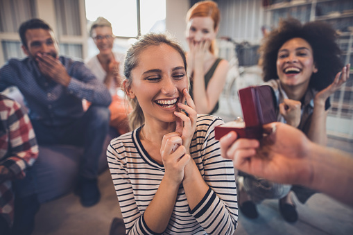 Surprised woman feeling excited while unrecognizable man is giving her an engagement ring while her friends are in the background.