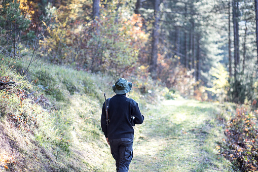 Rear  view of man in black clothes and green cap with rifle over his shoulder who is walking through the forest during sunny day