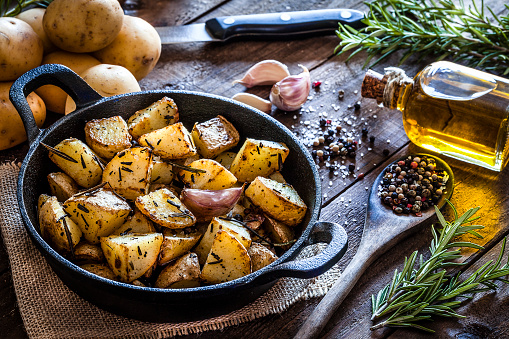 Cast iron pan filled with roasted potatoes shot on rustic wooden table. The cooking pan is at the left of an horizontal  frame and the ingredients for cooking the potatoes are all around the pan placed directly on the table. The ingredients includes are raw potatoes, rosemary, olive oil, salt, pepper and garlic. Predominant colors are brown and yellow. DSRL studio photo taken with Canon EOS 5D Mk II and Canon EF 100mm f/2.8L Macro IS USM