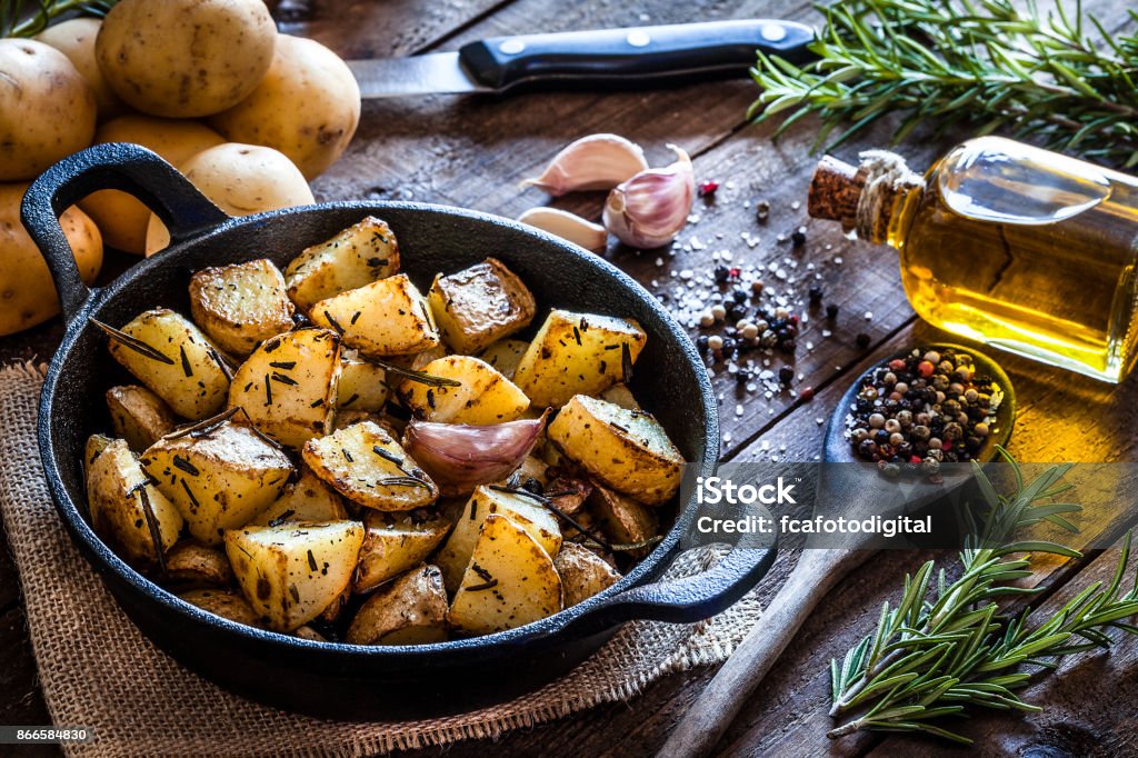 Roasted potatoes on wooden kitchen table Cast iron pan filled with roasted potatoes shot on rustic wooden table. The cooking pan is at the left of an horizontal  frame and the ingredients for cooking the potatoes are all around the pan placed directly on the table. The ingredients includes are raw potatoes, rosemary, olive oil, salt, pepper and garlic. Predominant colors are brown and yellow. DSRL studio photo taken with Canon EOS 5D Mk II and Canon EF 100mm f/2.8L Macro IS USM Raw Potato Stock Photo