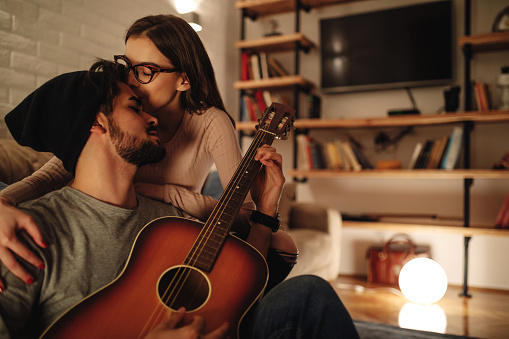 Shot of a young woman kissing her boyfriend while he's playing the guitar at home.