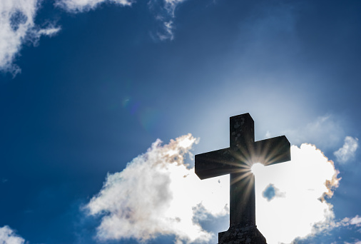 Silhouette of cross shape with sunrays and cloudy sky.