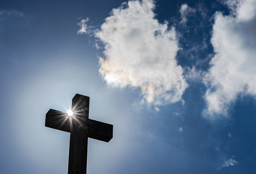Silhouette of cross shape with sunrays and cloudy sky.