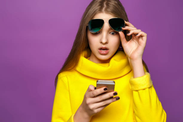 joven sorprendida retrato de mensaje en smartphone en el suéter amarillo brillantes, aislar sobre un fondo violeta - child surprise little girls human face fotografías e imágenes de stock