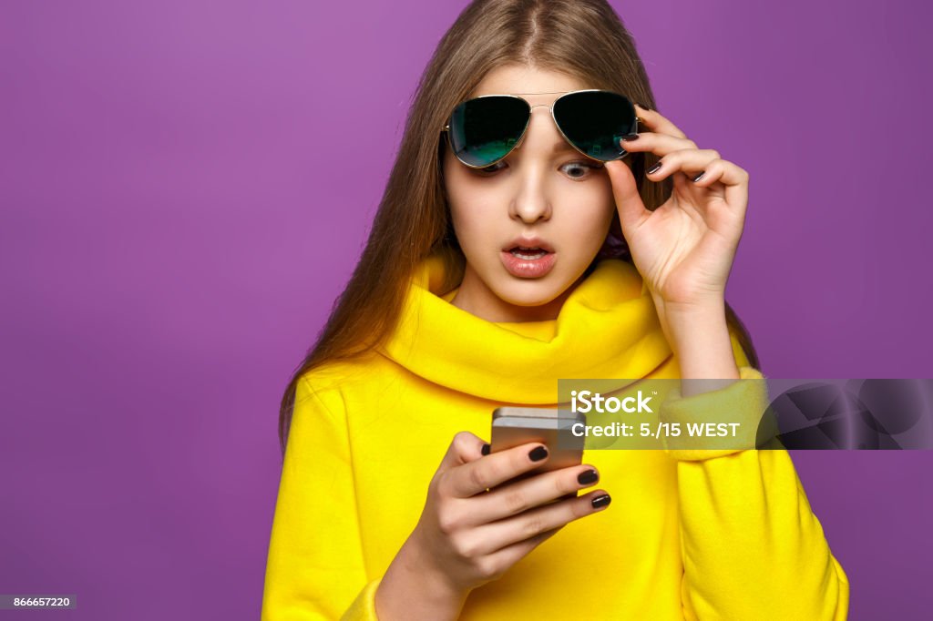 Portrait surprised young girl from message on smartphone in brightly yellow sweater, isolate on a violet background Surprise Stock Photo
