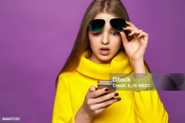 Portrait Surprised Young Girl From Message On Smartphone In Brightly Yellow Sweater Isolate On A Violet Background Stock Photo - Download Image Now