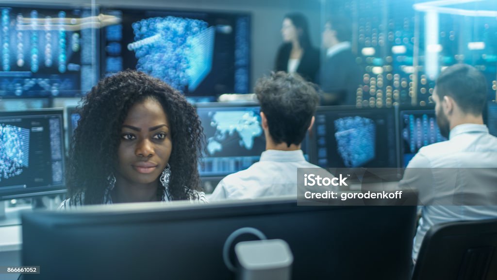 Female Computer Engineer Works on a Neural Network/ Artificial Intelligence Project with Her Multi-Ethnic Team of Specialist. Office Has Multiple Screens Showing 3D Visualization. Analyzing Stock Photo