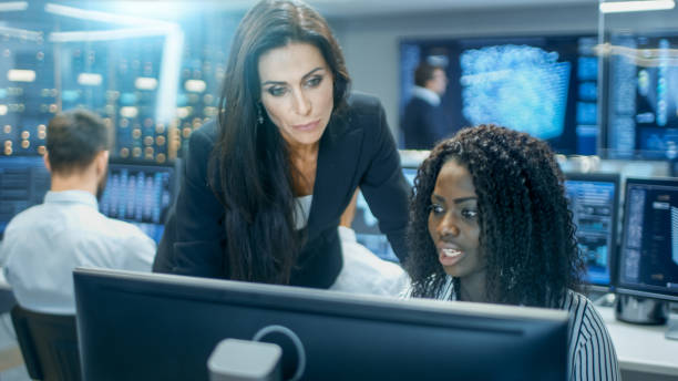 istock Female Team Leader Consults Young Computer Engineer. They Work in a Crowded Office on a Neural Network/ Artificial Intelligence Project. 866661062