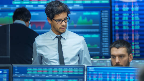 istock Stock Trader Inspects Others Work and Chat with Other. Busy Office with Multi-Ethnic Team of Brokers, Dealers. Displays Show Numbers and Infographics. 866661068
