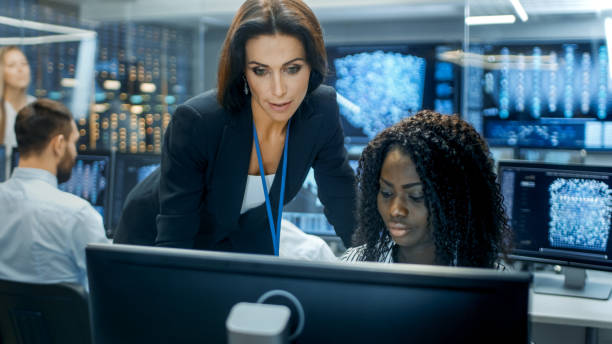 istock Female Team Leader Consults Young Computer Engineer. They Work in a Crowded Office on a Neural Network/ Artificial Intelligence Project. 866661070