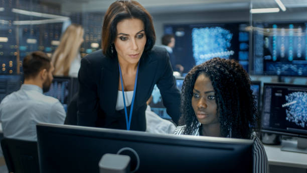istock Female Team Leader Consults Young Computer Engineer. They Work in a Crowded Office on a Neural Network/ Artificial Intelligence Project. 866661114