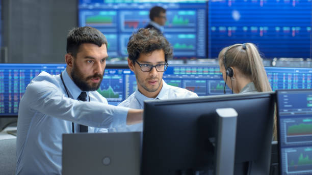 istock Professional Broker Consults Stock Exchange Trader at His Workstation. Multi-Ethnic Team at Stock Exchange Office is Busy Selling and Buying Stocks on the Market. Displays Show Relevant Data Numbers. 866662024