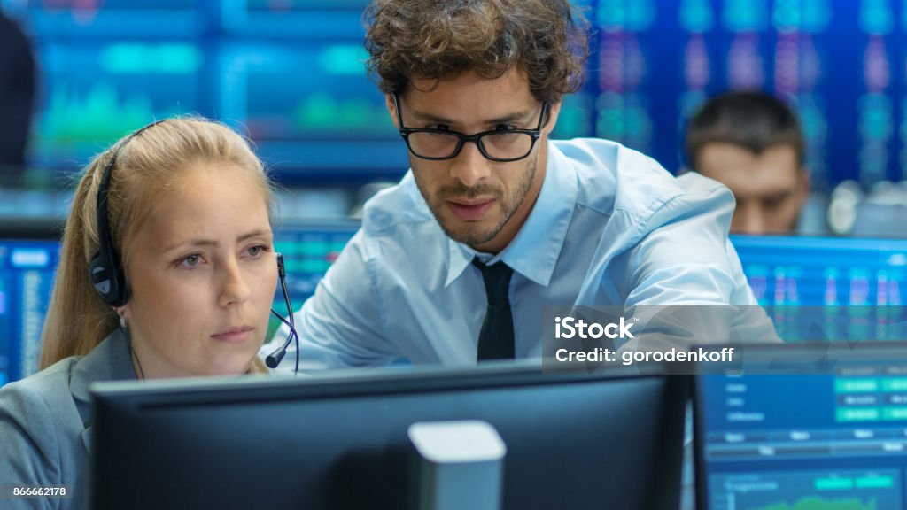 Experienced Stock Exchange Trader Teaches His Apprentice at Her Workstation. Multi-Ethnic Team at Stock Exchange Office is Busy Selling and Buying Stocks on the Market. Displays Show Relevant Data Numbers. Data Stock Photo