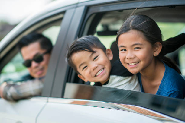 Road Trip An Asian family is on a road trip together. They are wearing casual clothes. The father is looking out the front window of a van, and the son and daughter are smiling out the back window. family-van-driving stock pictures, royalty-free photos & images