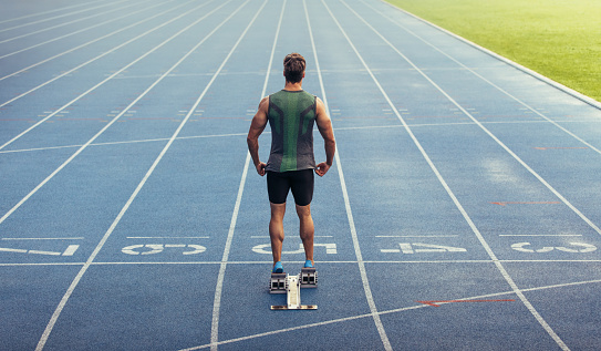 Rear view of an athlete ready to sprint on an all-weather running track. Runner using a starting block to start his run on race track.