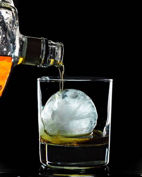 Round spherical ice cube ball in a cocktail glass with unique rim lighting against a black background, with an alcoholic beverage being poured into it. stock photo