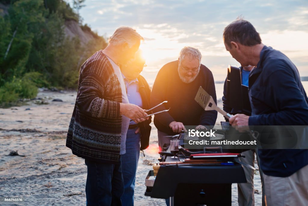 Five seniors brothers cooking fish on BBQ on the beach Five real seniors brothers, aged from 65 to 80, cooking their lunch on BBQ after a fishing day trip. Those guys sure know how to have fun! They are dressed in neutral-blue tones. Horizontal three-quarter length outdoors shot with some copy space and lense flare. This was taken in Quebec, Canada. Barbecue - Meal Stock Photo