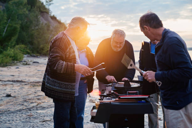 Five seniors brothers cooking fish on BBQ on the beach Five real seniors brothers, aged from 65 to 80, cooking their lunch on BBQ after a fishing day trip. Those guys sure know how to have fun! They are dressed in neutral-blue tones. Horizontal three-quarter length outdoors shot with some copy space and lense flare. This was taken in Quebec, Canada. family bbq beach stock pictures, royalty-free photos & images