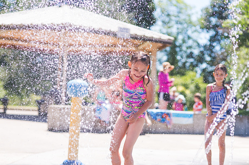 A group of young children frolic and run in a playground splash pad. It is a sunny, perfect day for getting wet and playing hard! A young girl in a swimsuit is running through the sprinklers and yelling and laughing as the water droplets cascade all around and her friends wait in the background.